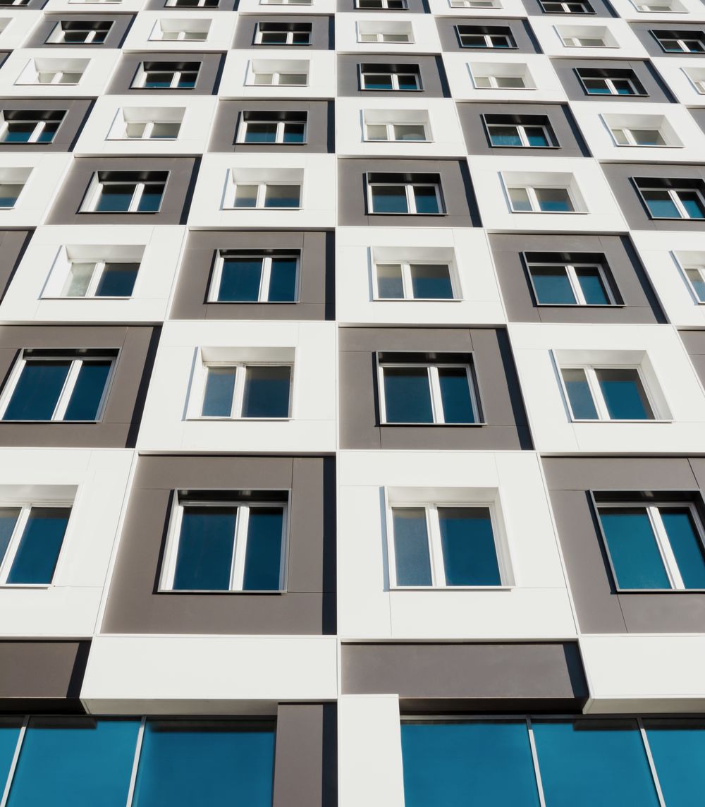 Façade d’un immeuble de logements contemporain vue en contre-plongée, fenêtres encadrées de blanc et de gris qui se répètent jusqu’au ciel.